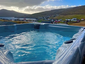 A hot tub with a view of a small village and hills in the background. - Modern 4BR Luxury Home with Ocean View & Jacuzzi (Sandavágur)