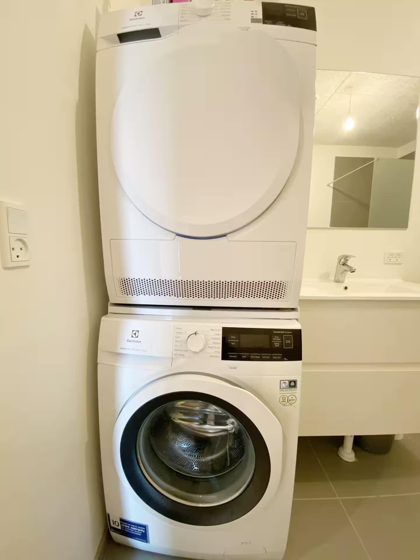 A front-loading washing machine and dryer stacked in a small laundry room with a sink and mirror.