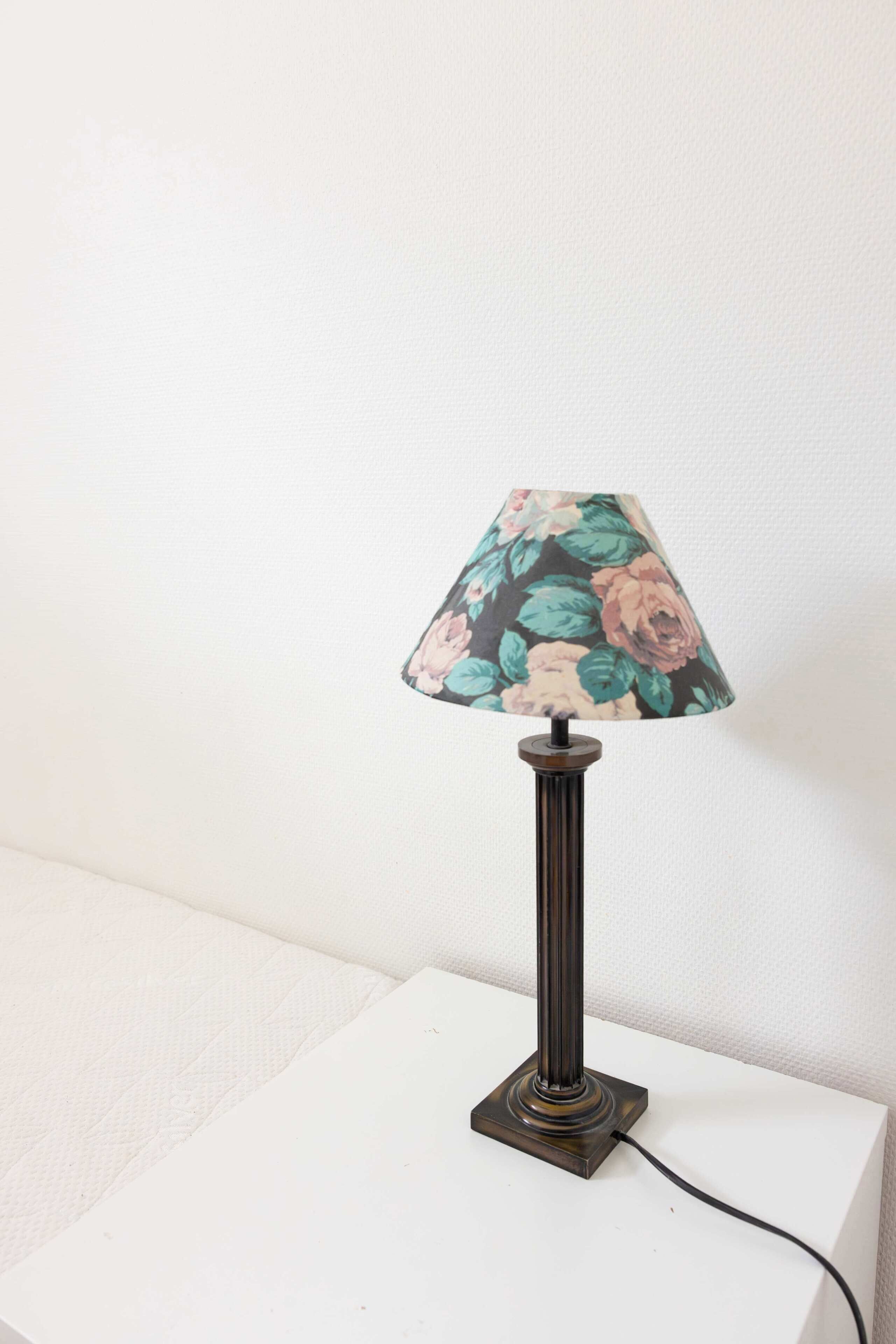 Une lampe de table avec un abat-jour à motifs floraux, posée sur une table blanche devant un mur uni.