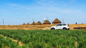 Een witte SUV geparkeerd in een veld met groene gewassen, omringd door traditionele huizen met rieten daken en een heldere blauwe lucht.