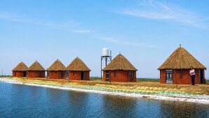 Een rij rieten hutten aan een waterlichaam, met een watertoren en een persoon die voor de laatste hut staat.