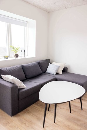 Ein modernes Wohnzimmer mit einem grauen Ecksofa, einem weißen runden Tisch und einem Fenster mit einem Blumentopf auf der Fensterbank.