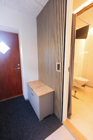 Huoneessa on puinen työpöytä, pystysuorista lankuista tehty seinä, ovi sekä kylpyhuone, jossa on wc ja pesuallas.