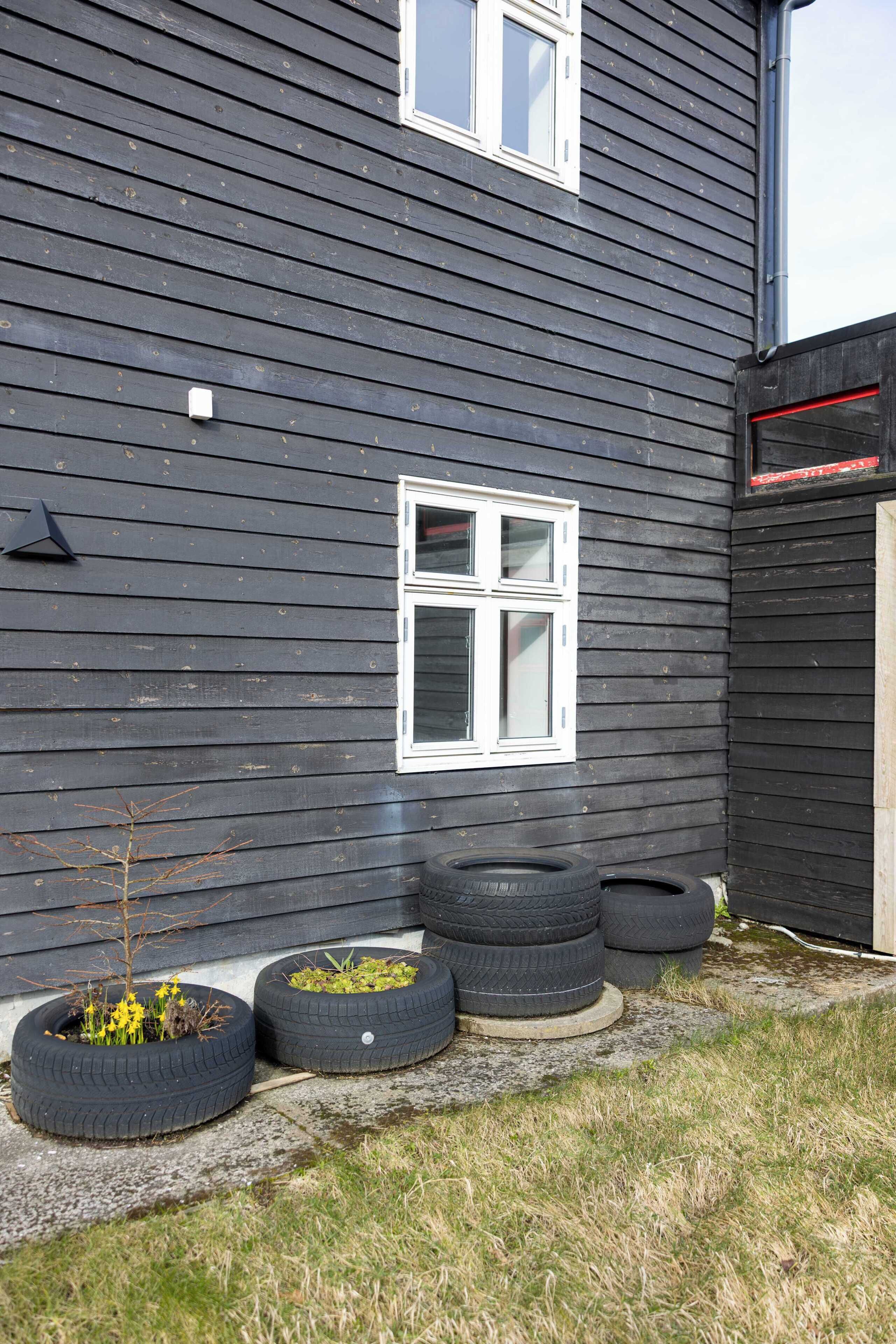 Una casa con fachada oscura, una ventana blanca y un pequeño jardín con flores amarillas y macetas hechas con neumáticos.