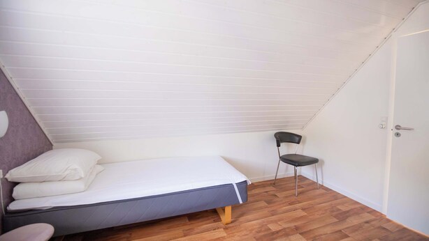 A small, single-bed room with a sloped ceiling, a wooden floor, a chair, and a door.