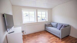 A room with a grey sofa, a white desk, and a television on the left. There are two windows with a view of houses outside.