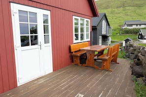 A wooden deck with a bench and table, a red building with white trim, and a view of a green landscape. - Summerhouse in Gjogv At the Edge of the Faroes (Gjógv)