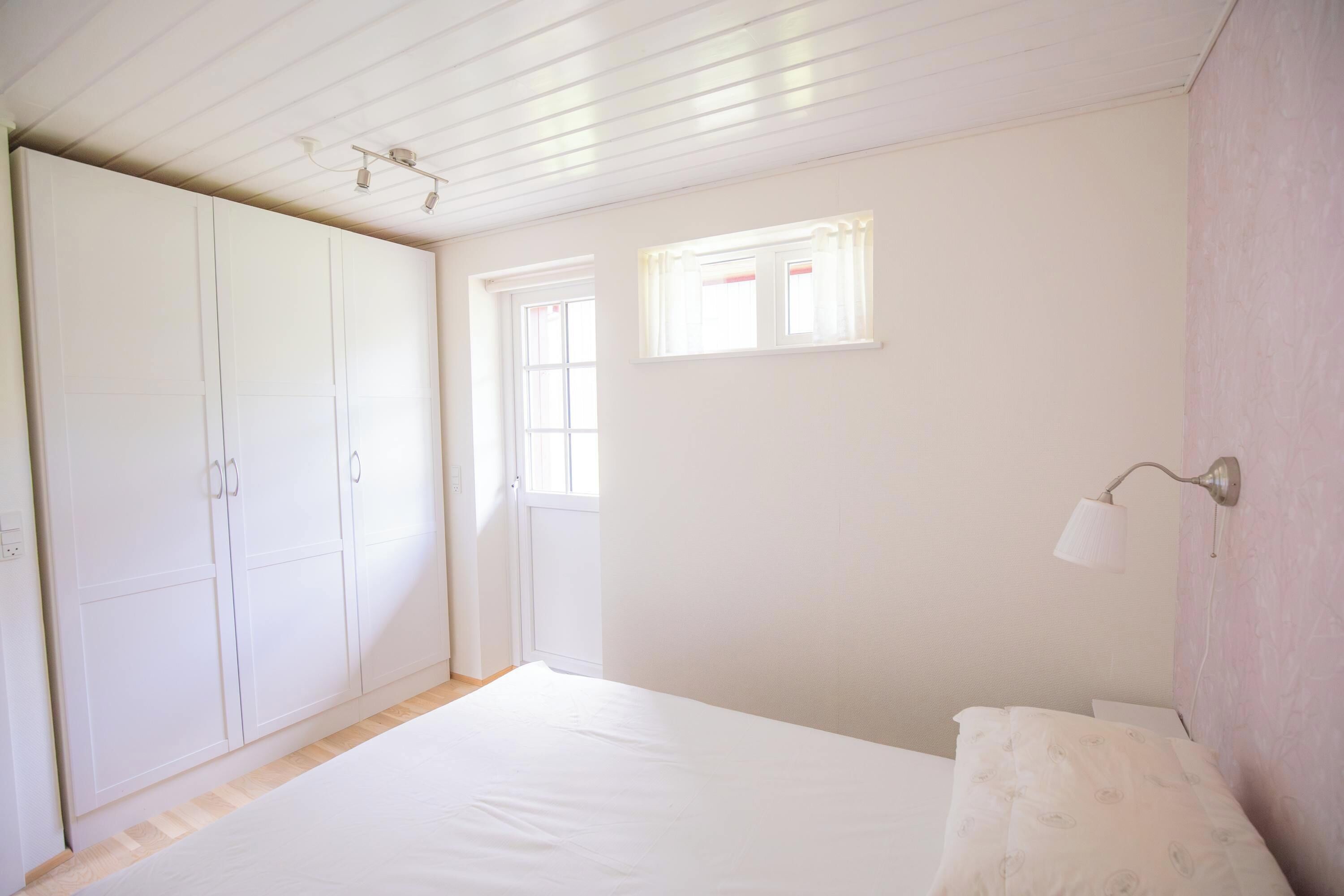 A bedroom with a bed, a wardrobe, a window, and a wall-mounted lamp.