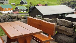 A wooden picnic table with benches on a wooden deck, overlooking a green hillside with houses.