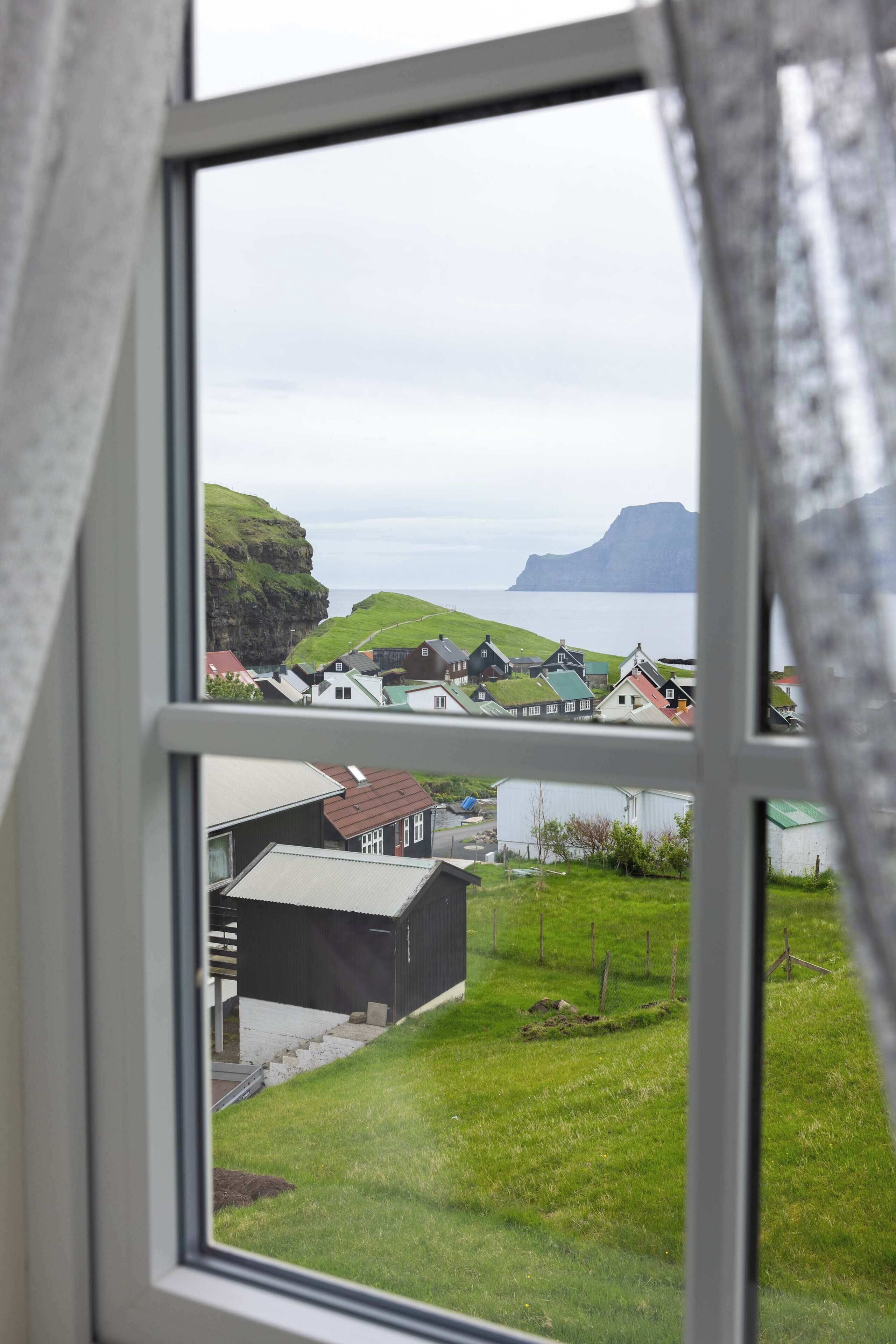 A window view of a coastal village with houses, a grassy area, and a cliff.