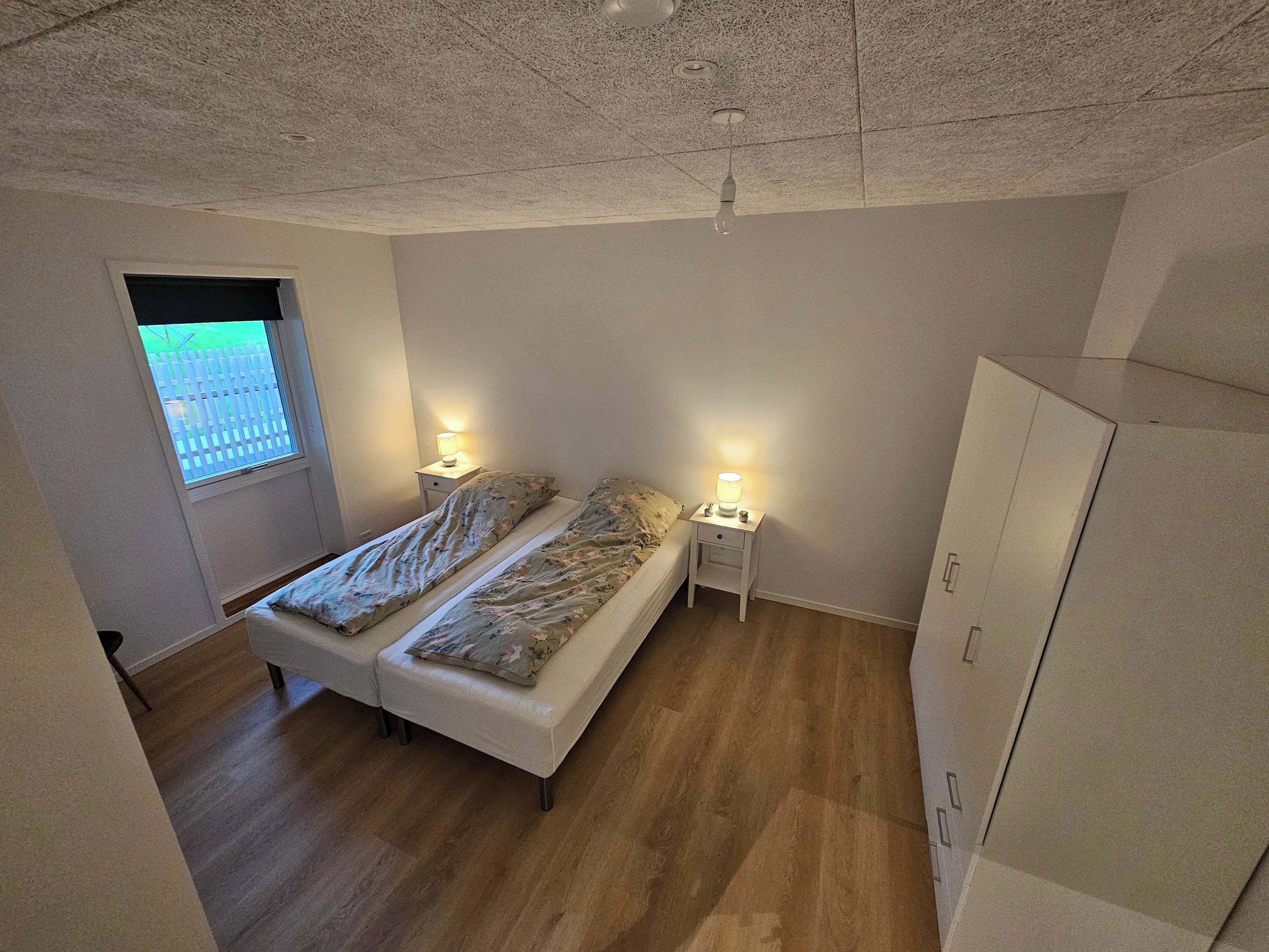 Une chambre à coucher avec un lit double, deux tables de chevet avec des lampes, une armoire et une fenêtre donnant sur une clôture.
