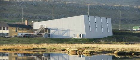 A modern building with a flat roof and a large reflective surface, situated next to a body of water and a grassy area.