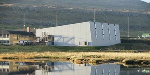 A modern building with a flat roof and a large reflective surface, situated next to a body of water and a grassy area. - Secluded Home with Mountain Views in Skalavik (Skálavík)