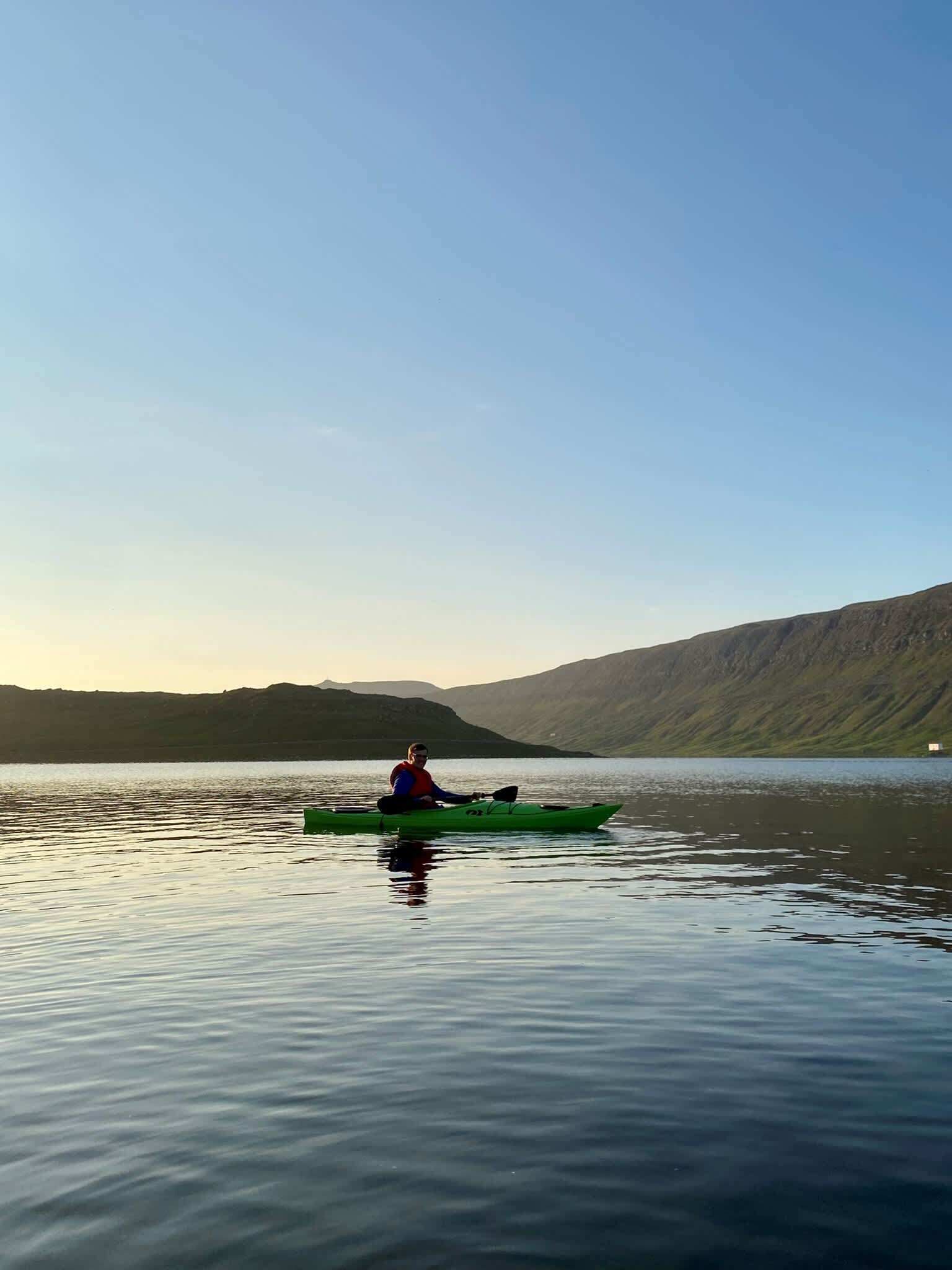 Eine Person paddelt mit einem Kajak auf ruhigem Wasser, im Hintergrund eine hügelige Landschaft.