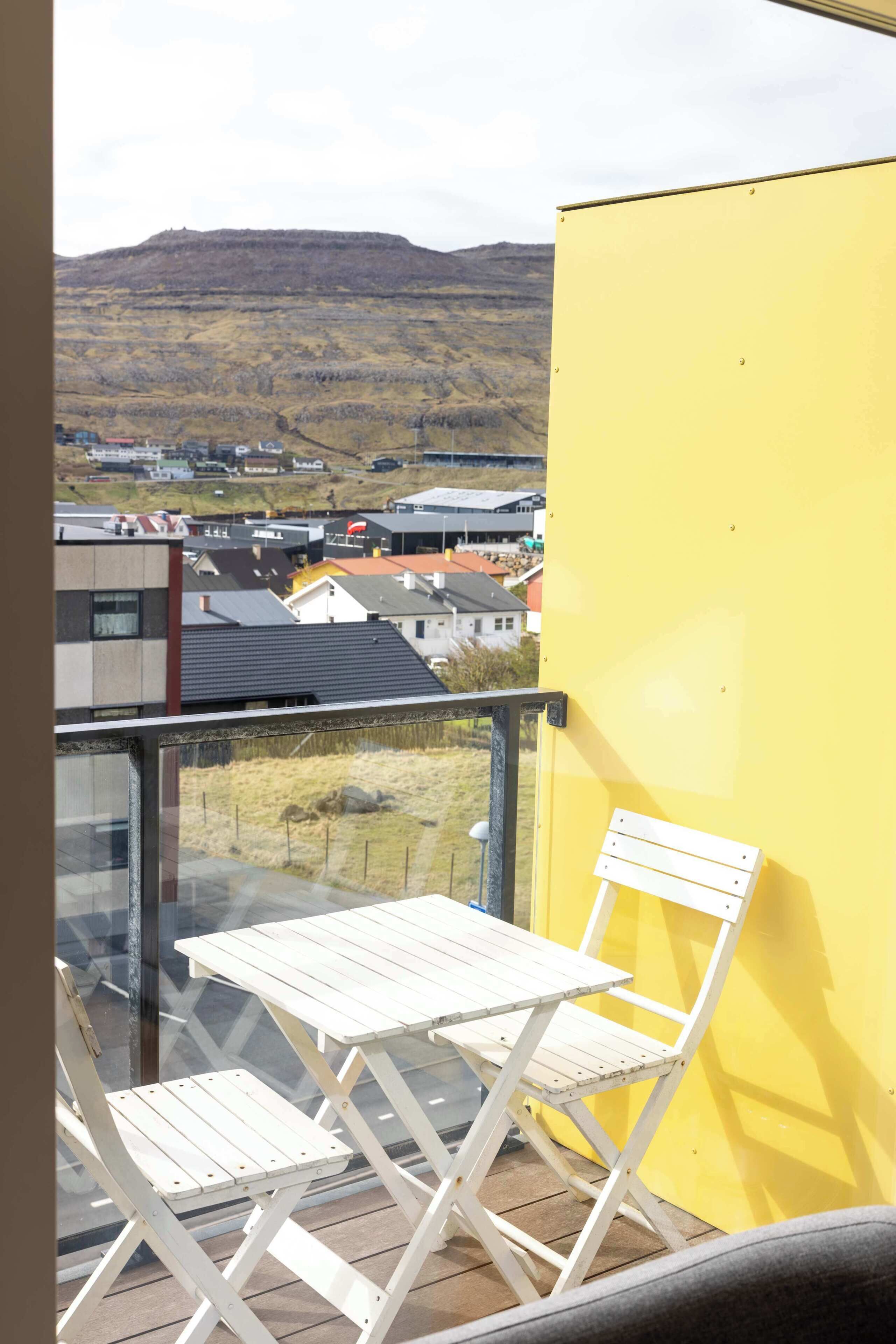 Un balcone con un tavolo e una sedia pieghevoli bianchi, vista su una zona residenziale con case e una collina sullo sfondo.