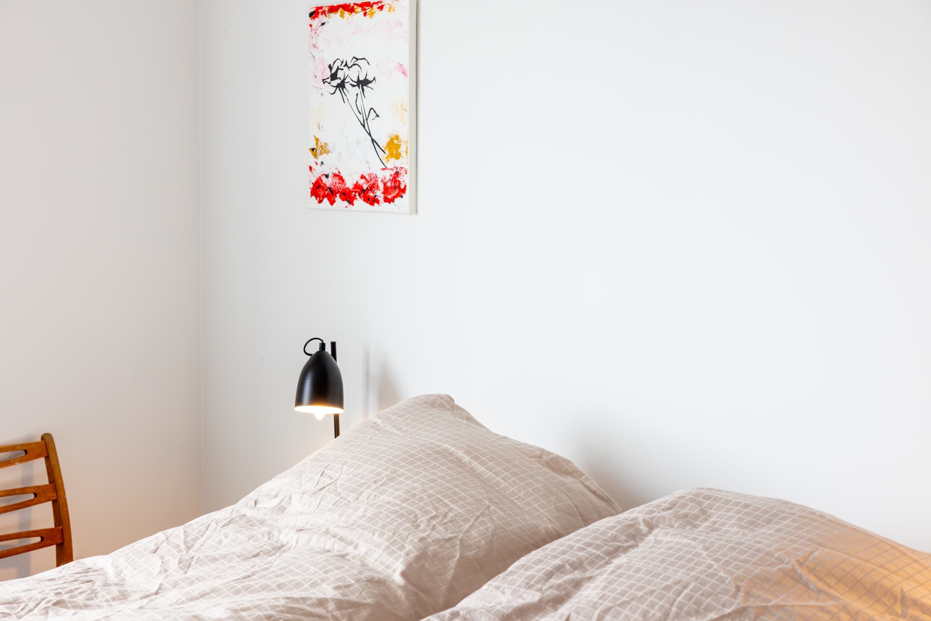 A bedroom with a bed, a chair, a wall-mounted lamp, and a framed artwork.