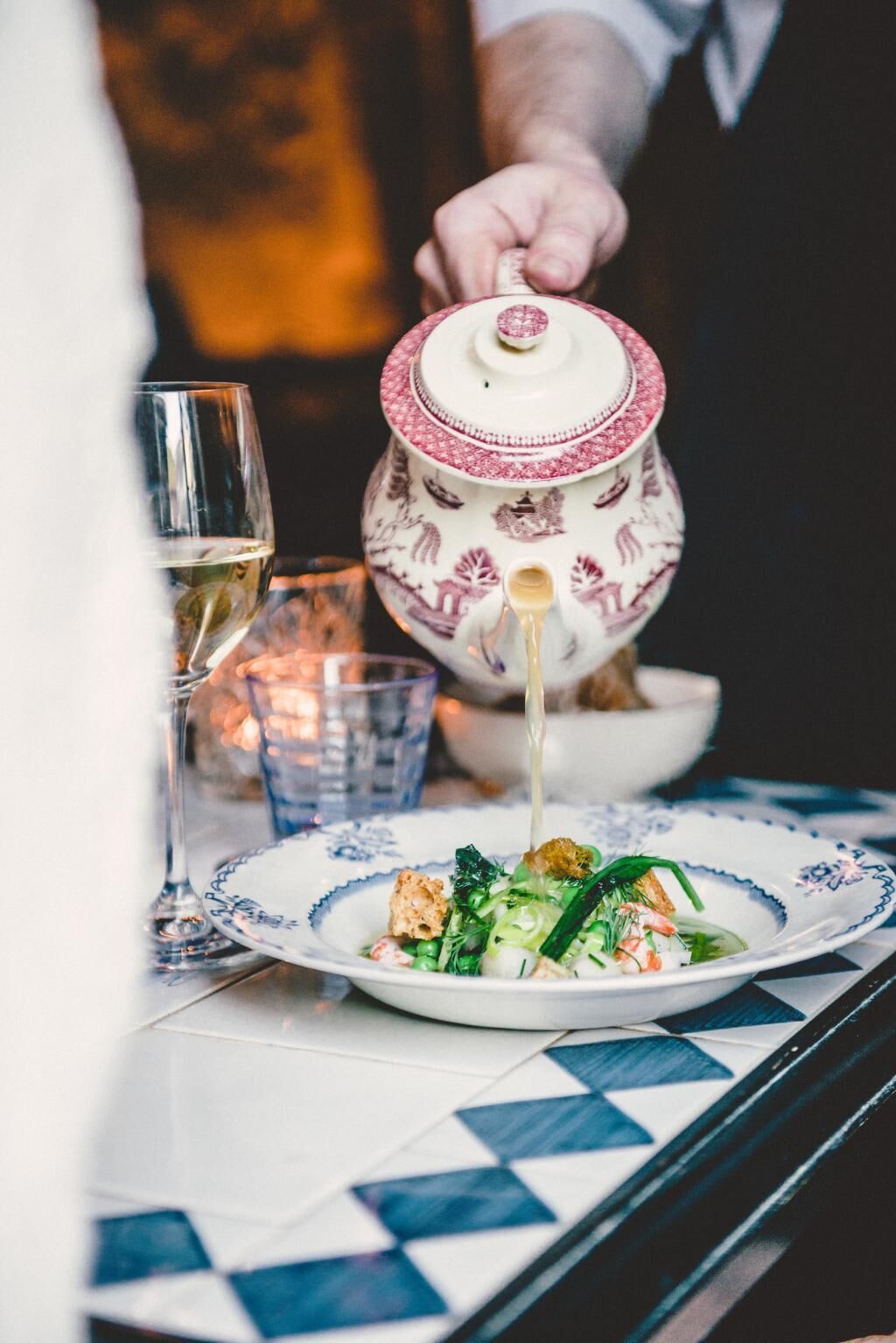 A dining table with a plate of food, a teapot pouring liquid, and glasses of a clear liquid.