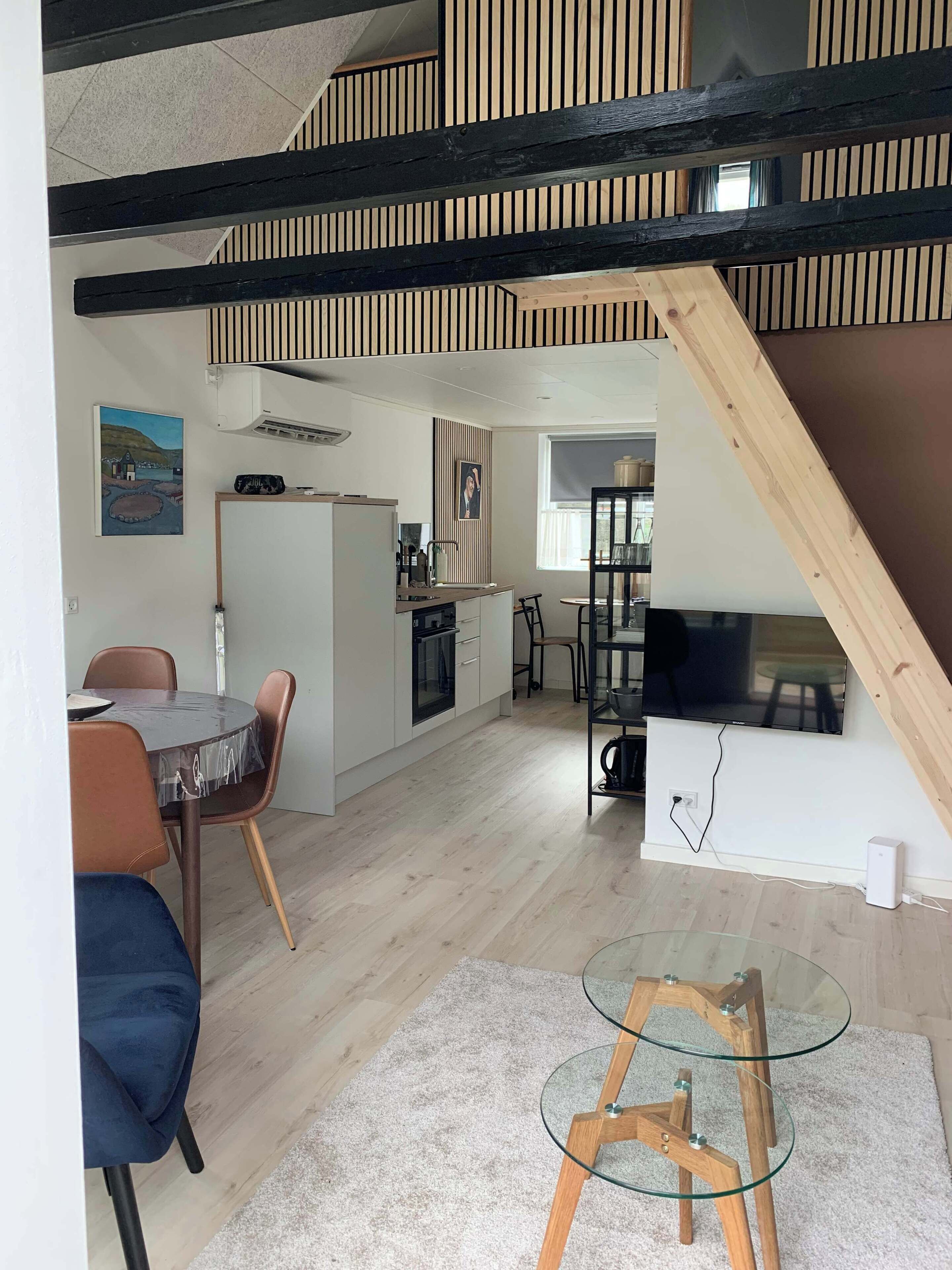 Une cuisine moderne avec des meubles blancs, un coin repas doté d’une table ronde et de chaises, et un espace en mezzanine avec un escalier en bois.