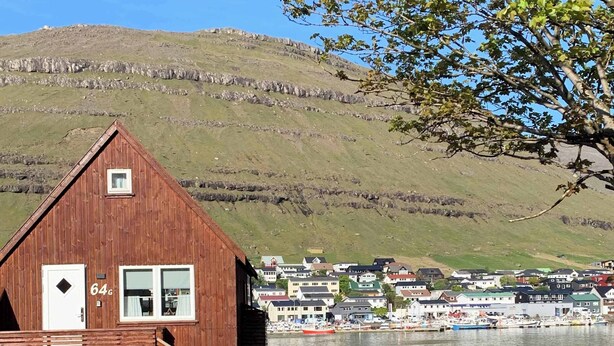 Eine Holzhütte mit kleinem Fenster und Tür, umgeben von viel Grün und Blick auf einen Hafen mit Booten.