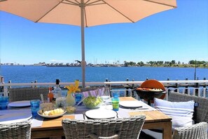 Un espacio para comer al aire libre con vista al agua, mobiliario de mimbre y una mesa dispuesta para una comida.