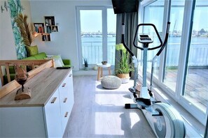 A modern living room with a view of the water, a wooden bench, a small table, and a stationary bike. - Holiday home in Kappeln with swimming pool (Kappeln)