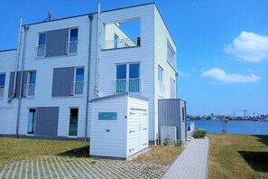 Un edificio de varios pisos con techo plano, una estructura pequeña adosada y un camino pavimentado que lleva a una masa de agua.