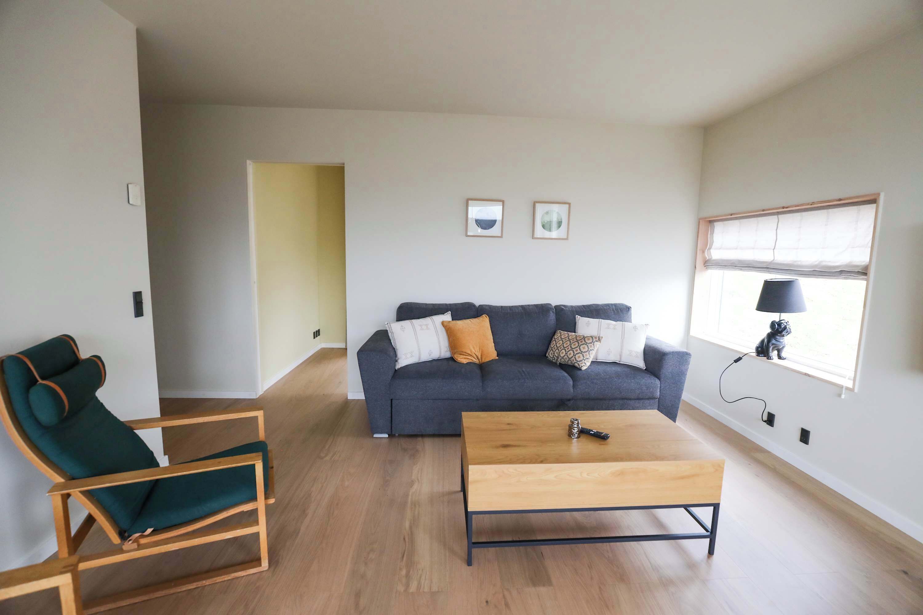 Een woonkamer met een grijze bank, een houten salontafel en een groene fauteuil. Twee ingekaderde foto's hangen aan de muur.