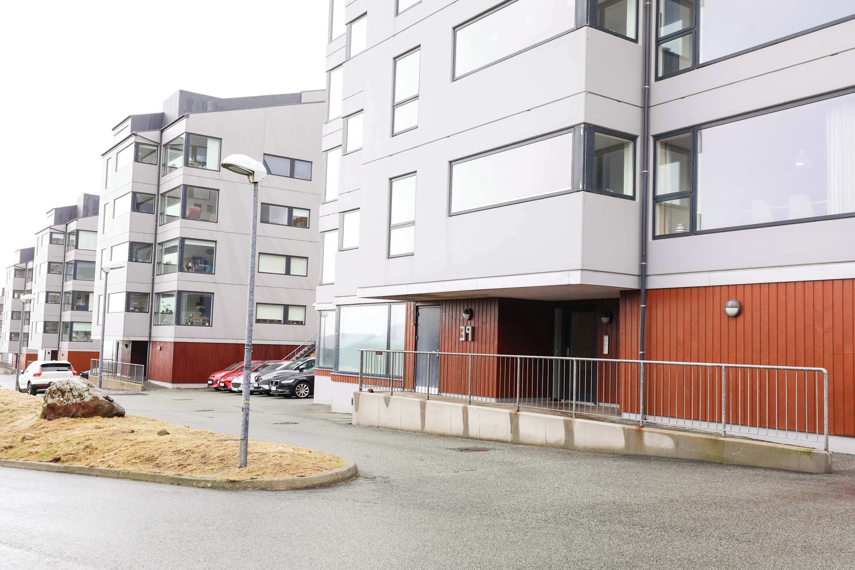 Ein mehrgeschossiges Wohngebäude mit roter und weißer Fassade, einem sichtbaren Eingang mit Metallgeländer und parkenden Autos im Vordergrund.