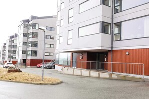A multi-story residential building with a red and white facade, a visible entrance with a metal railing, and parked cars in the foreground.
