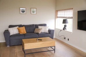 A living room with a grey sofa, a wooden coffee table, a TV mounted on the wall, and a window with a lamp on the sill.