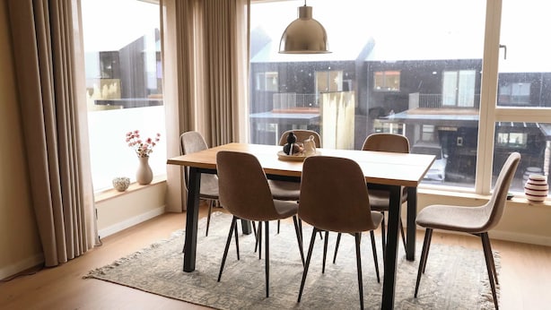 Un espace repas comprenant une table et des chaises, un luminaire suspendu et une vue sur les bâtiments à travers de grandes fenêtres.