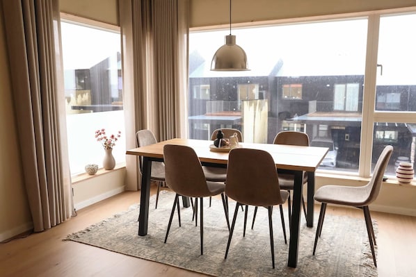 A dining area with a table and chairs, a hanging light fixture, and a view of buildings through large windows. - Light, Calm and Modern Corner Apartment (Argir)