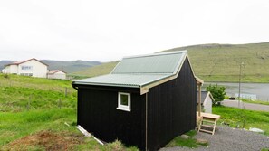 Un petit bâtiment en bois noir, doté d’un toit en pente, situé sur une colline herbeuse, avec un plan d’eau et des collines en arrière-plan.