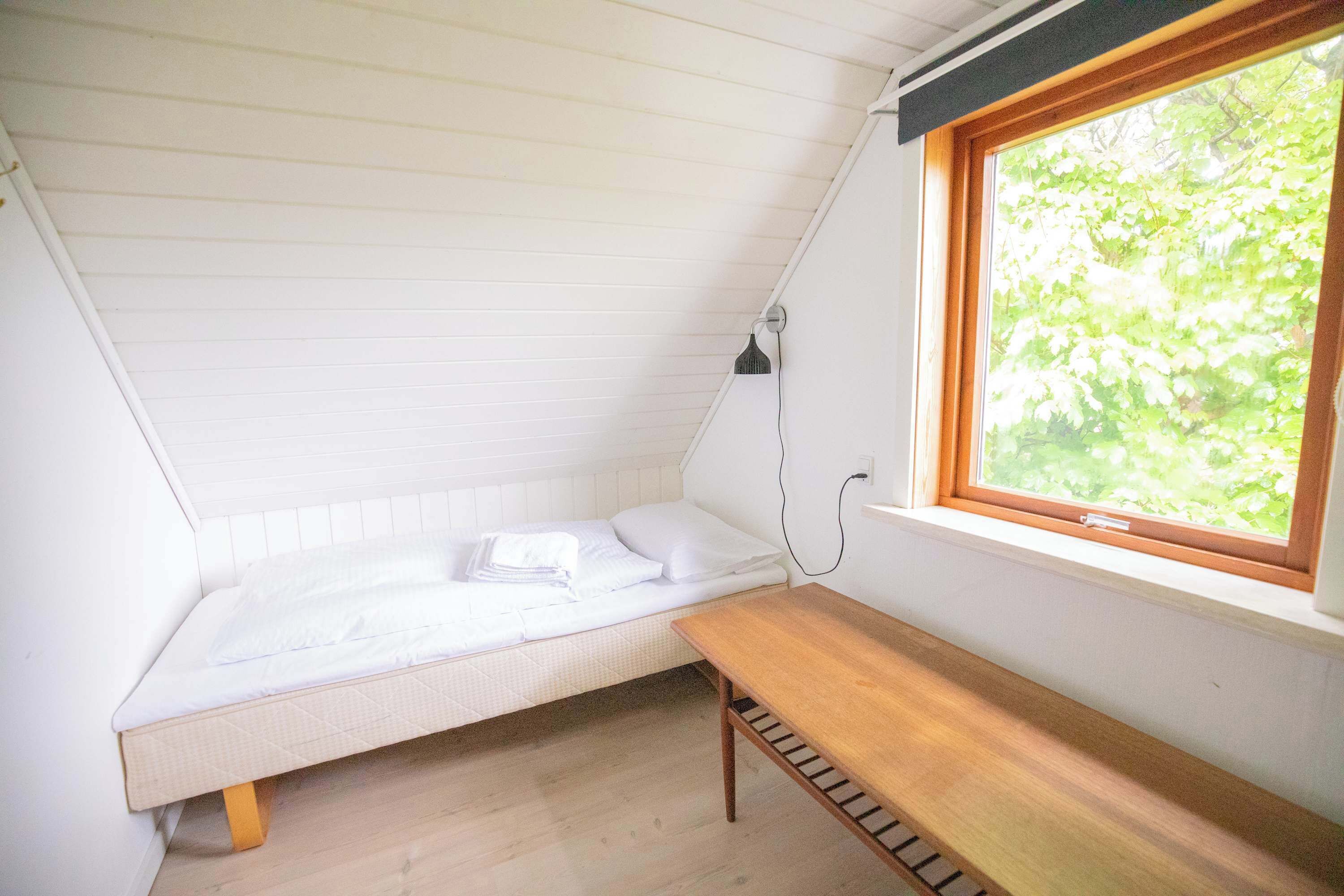 Ein kleiner, schräger Raum mit einem Bett, einer Sitzbank und einem Fenster mit Blick auf Grünflächen.