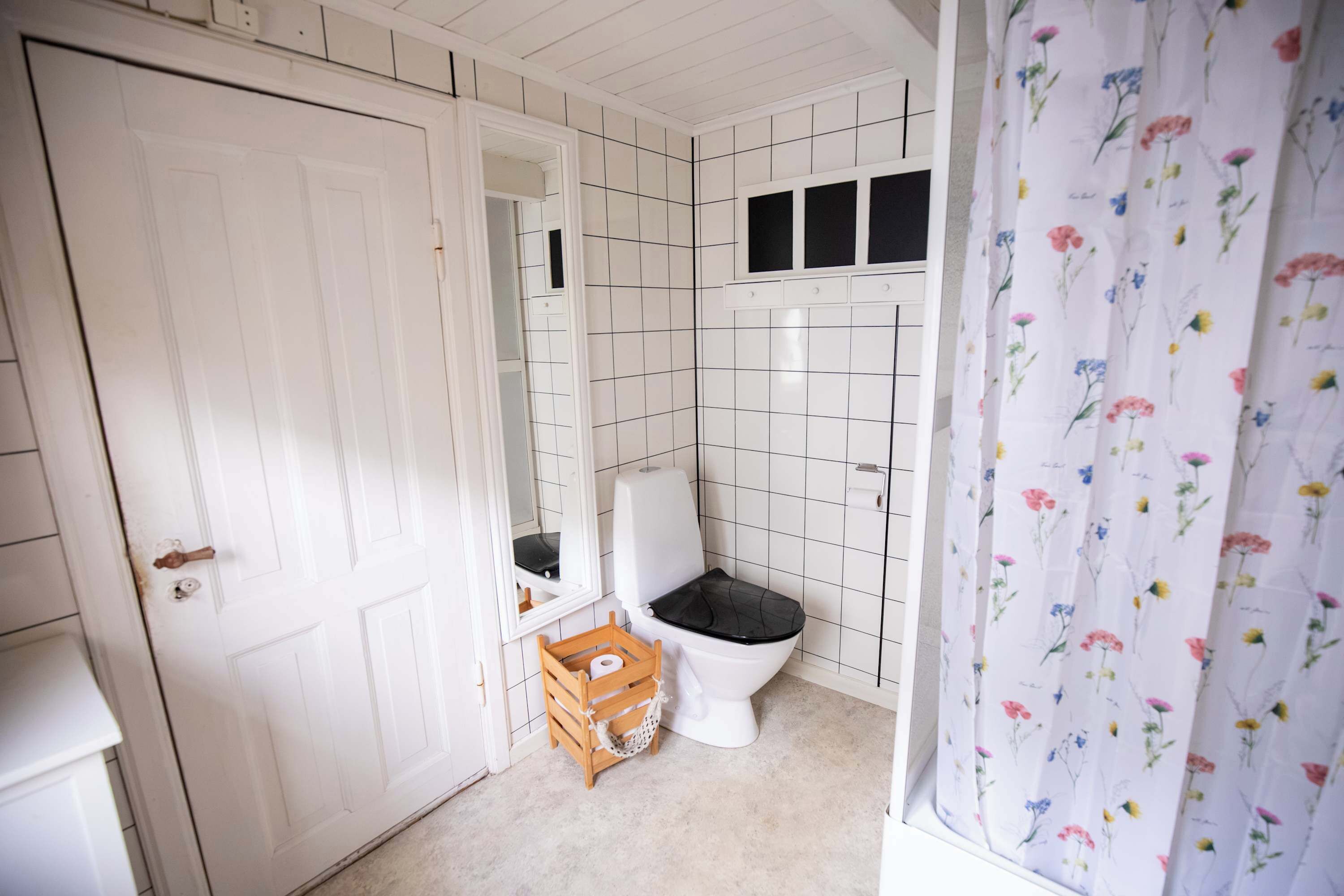 Un baño con paredes de azulejos blancos, un inodoro blanco, un caja de madera con un rollo de papel higiénico y una cortina de ducha estampada con motivos florales.
