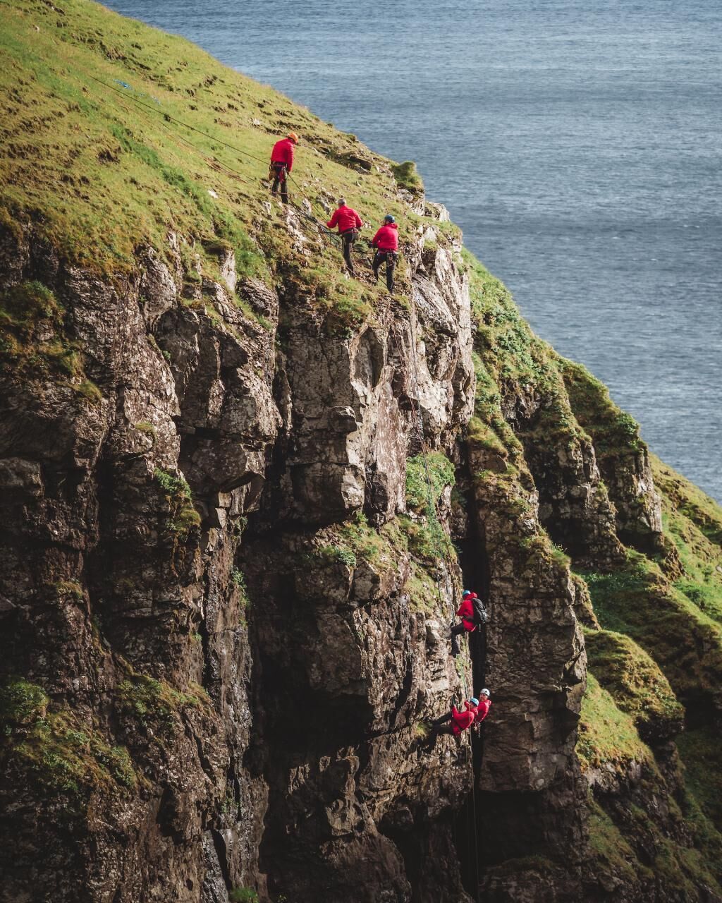 A group of people in red jackets climbing a rocky cliff by the sea.