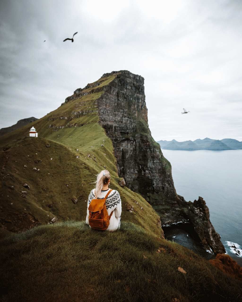 A person sitting on a grassy hill with a cliff and a small white building in the background.