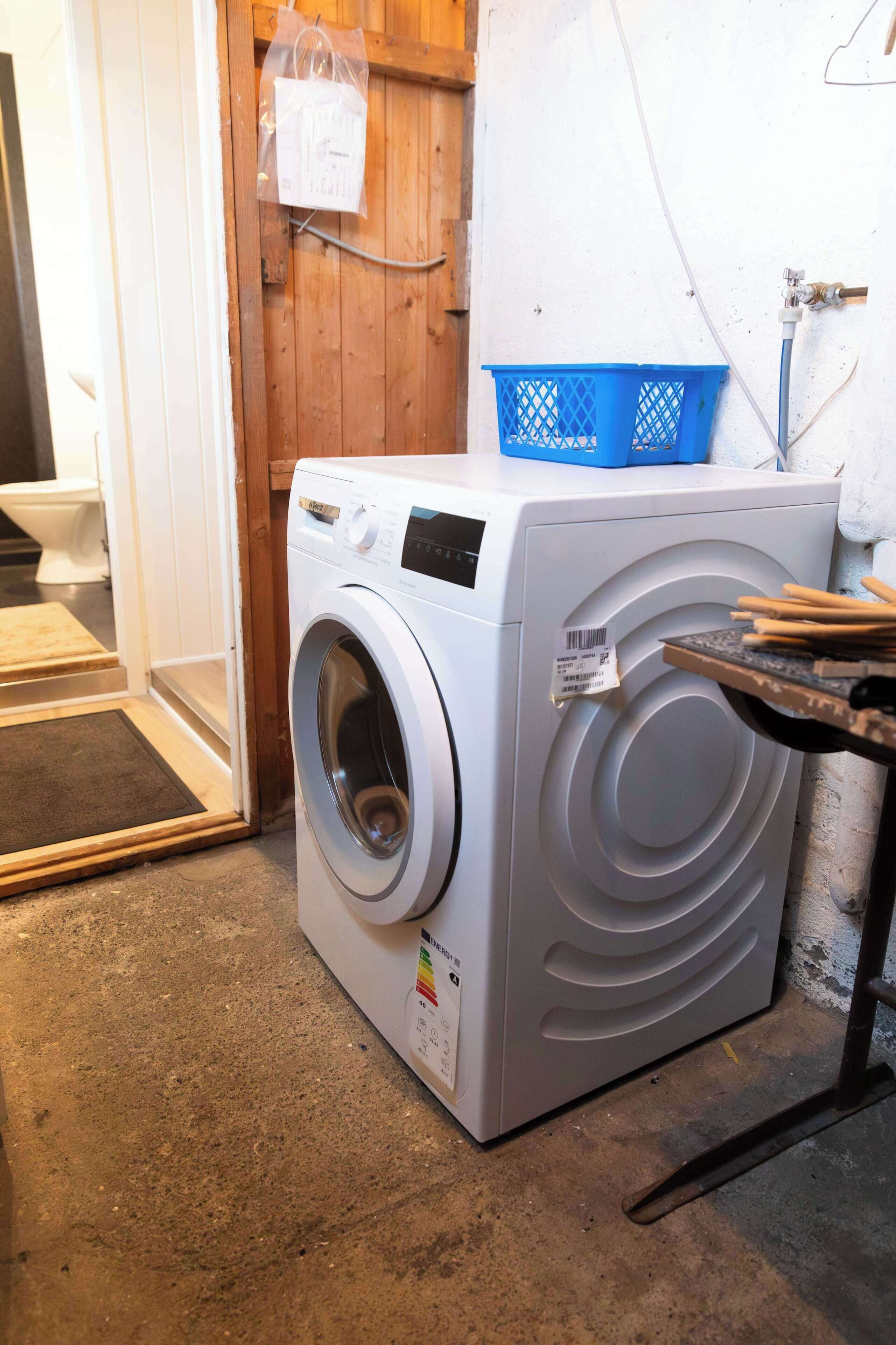 A front-loading washing machine in a laundry area with a wooden door, a blue laundry basket, and a white wall.