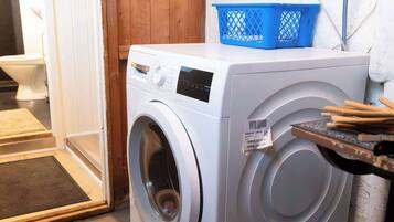 A front-loading washing machine in a laundry area with a wooden door, a blue laundry basket, and a white wall.