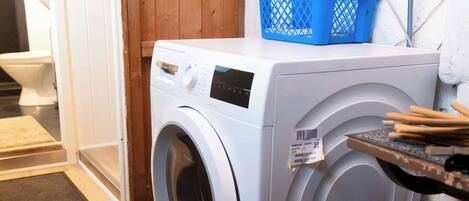 Un lave-linge à chargement frontal dans un espace buanderie, avec une porte en bois, un panier à linge bleu et un mur blanc.