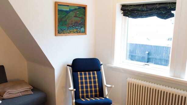 Ein Wohnzimmer mit blauem Sessel, einem kleinen Holztisch, einer Couch, einem Gemälde an der Wand und einem Fenster mit Blick.