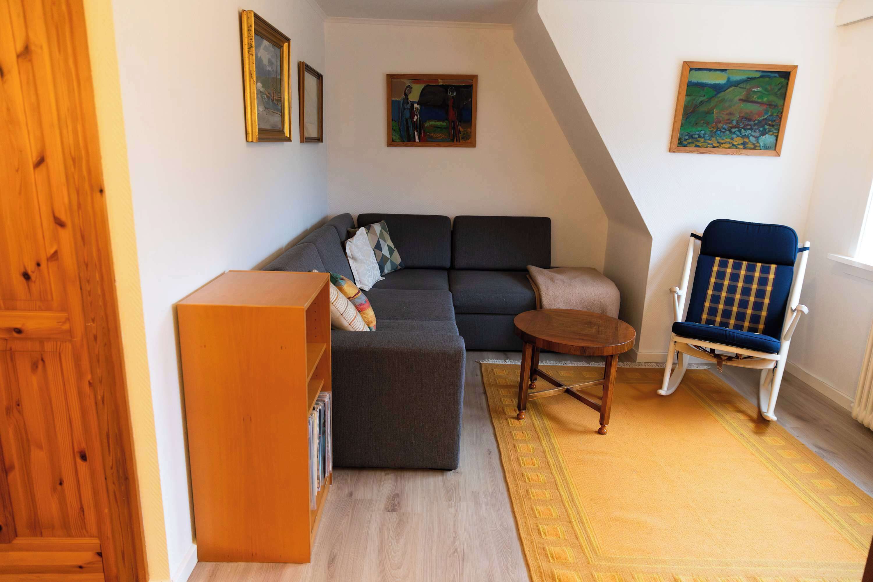 Ein gemütliches Wohnzimmer mit einer Eckcouch, einem kleinen runden Tisch, einem Schaukelstuhl und einem Bücherregal.