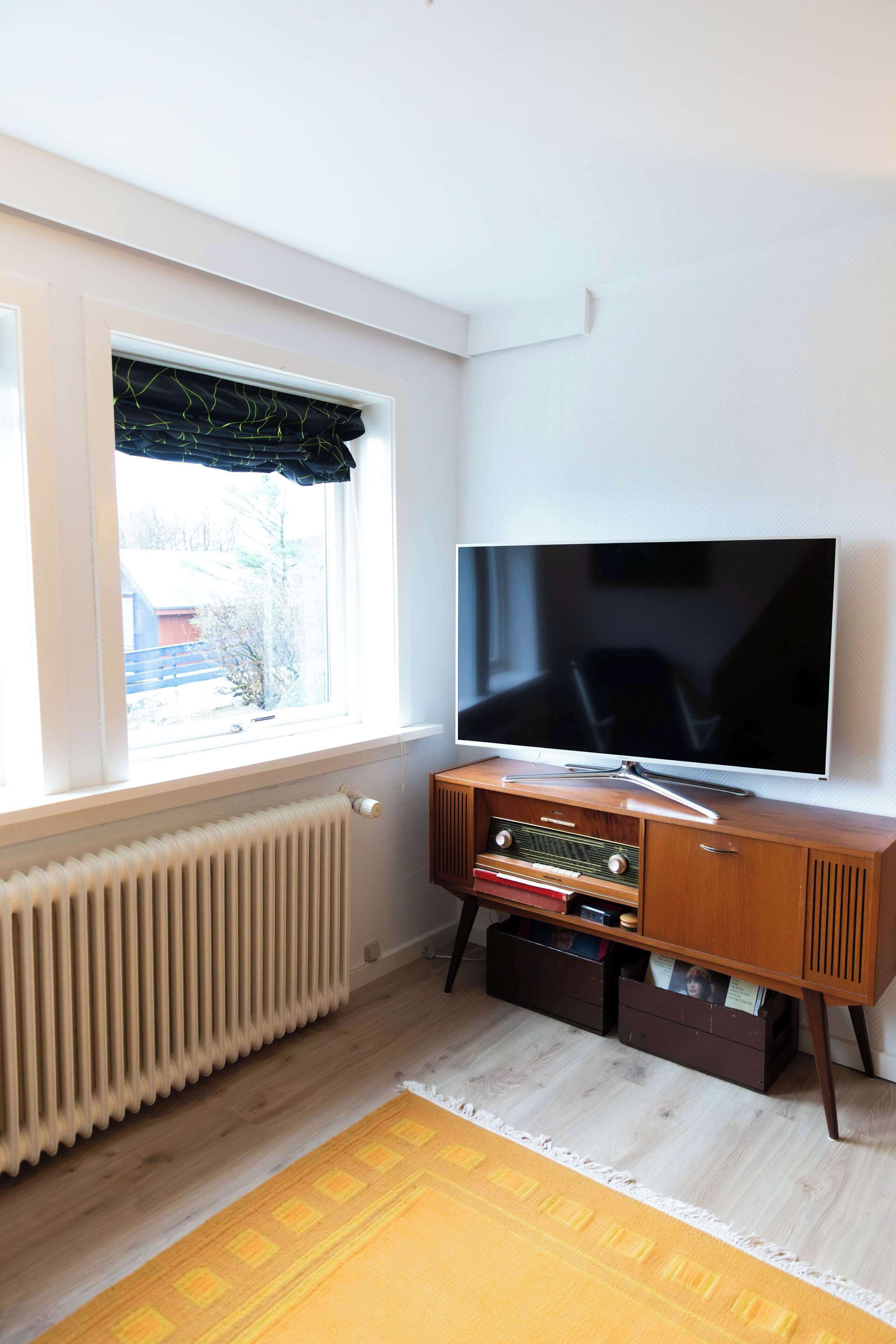 Una habitación con una televisión de pantalla grande sobre un soporte de madera, una ventana con cortina negra, un radiador y una alfombra estampada.