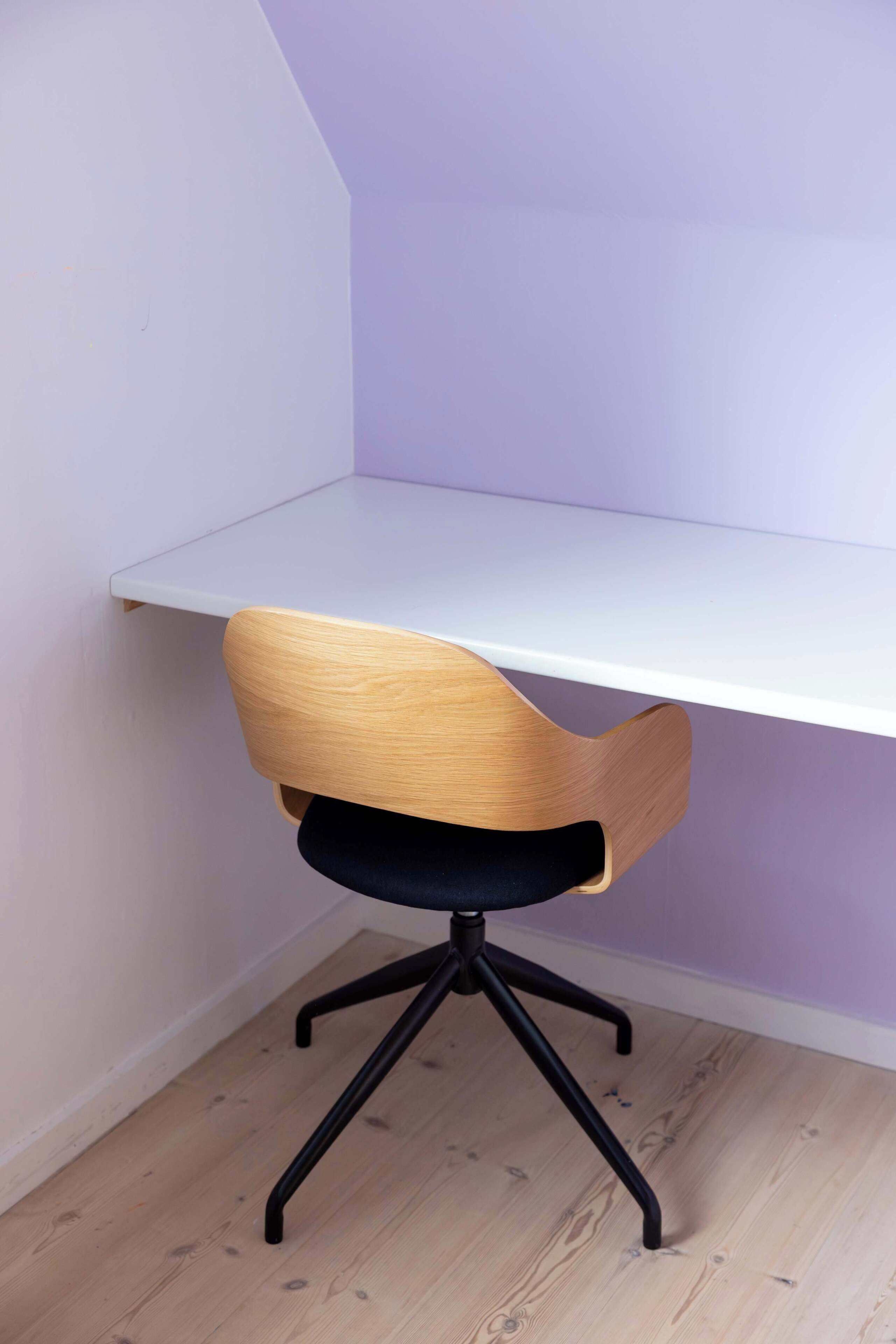 Ein Stuhl mit Holzlehne und schwarzer Sitzfläche, vor einem weißen Schreibtisch an einer violetten Wand platziert.