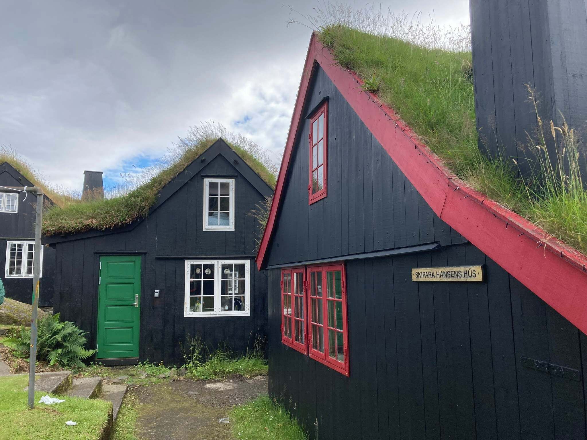 Ein traditionelles, schwarzes Holzhaus mit rotem Dach und grüner Tür, mit Gründächern und einem Schild, auf dem 'Skipara Hansens Hus' steht.