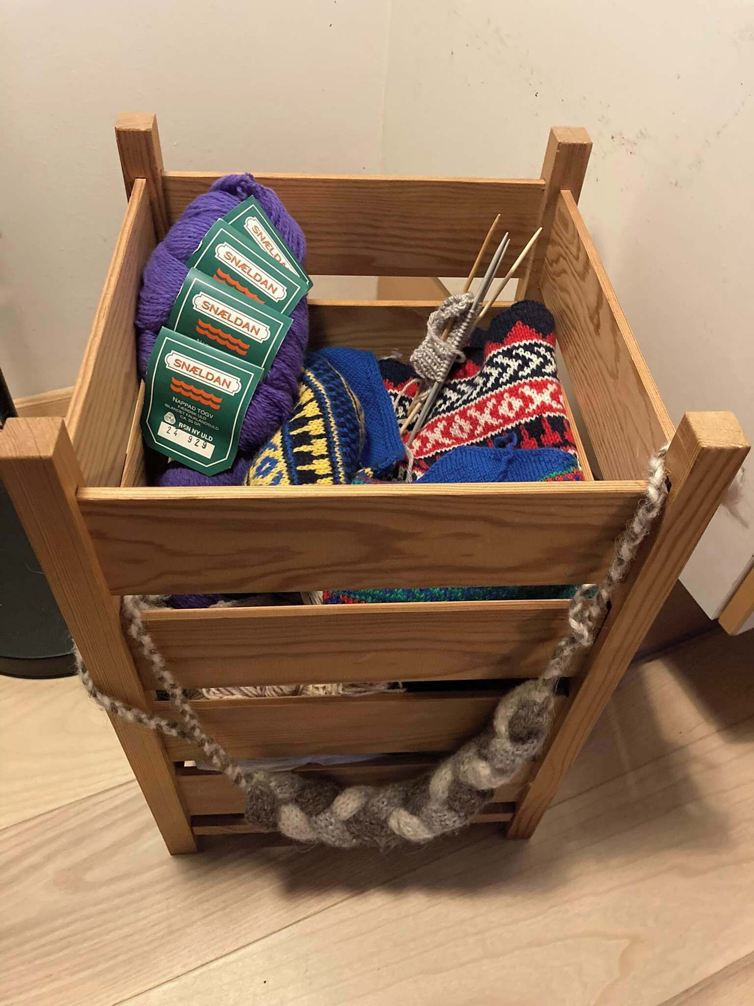 A wooden crate with a handle, containing various items including a purple knitted item, a multicolored knitted item, and a green package labeled 'SNELDAN'.