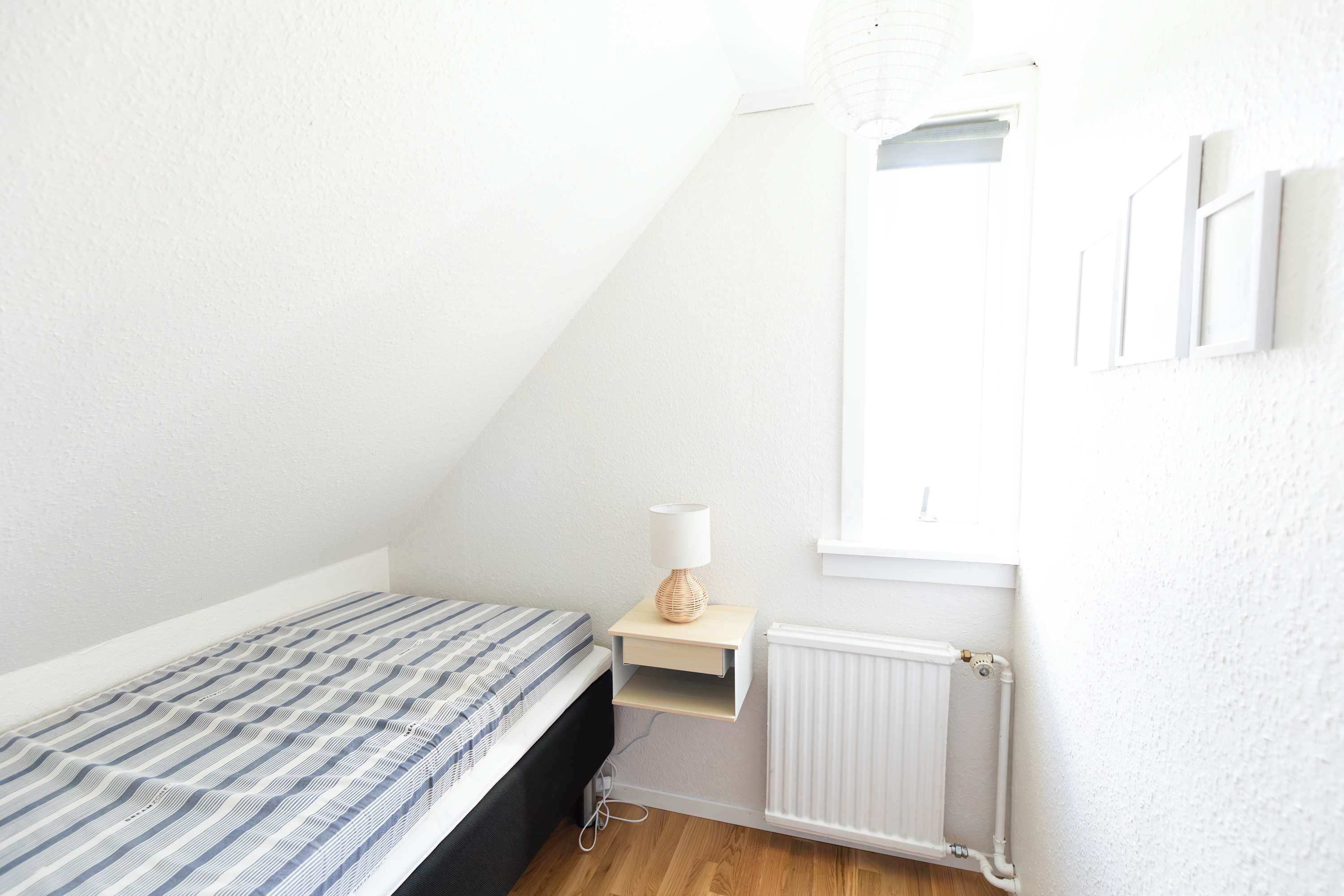 A small, sparsely furnished room with a bed, a nightstand, a lamp, and a radiator.