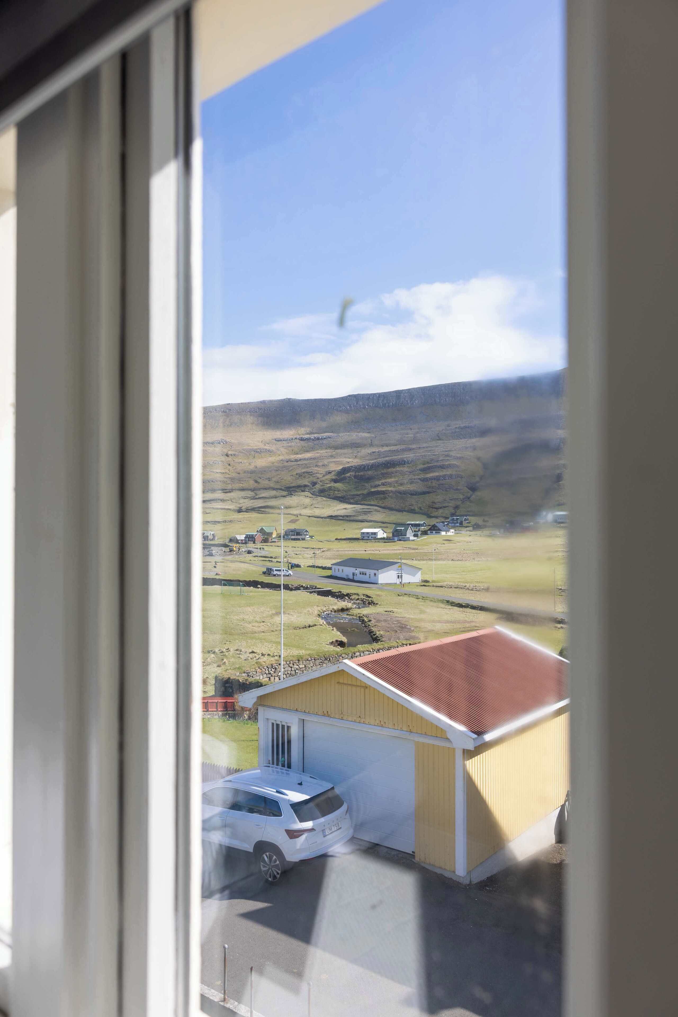 Vista dalla finestra di un edificio, con un'auto parcheggiata di fronte, un piccolo edificio con tetto rosso e un paesaggio collinare sullo sfondo.