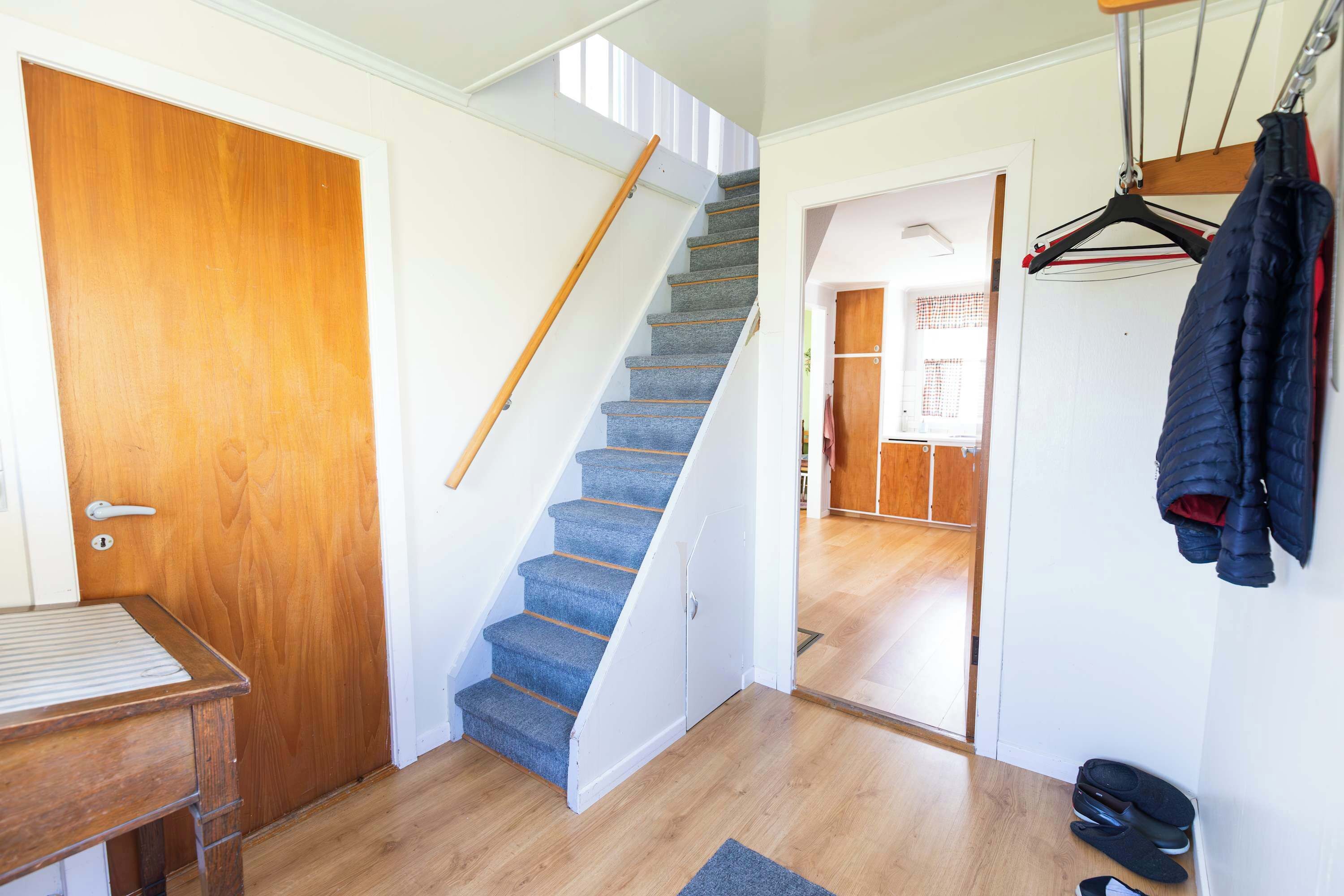 Una escalera que conduce a un segundo piso, una puerta de madera, un perchero con chaquetas y un par de zapatos en el suelo.