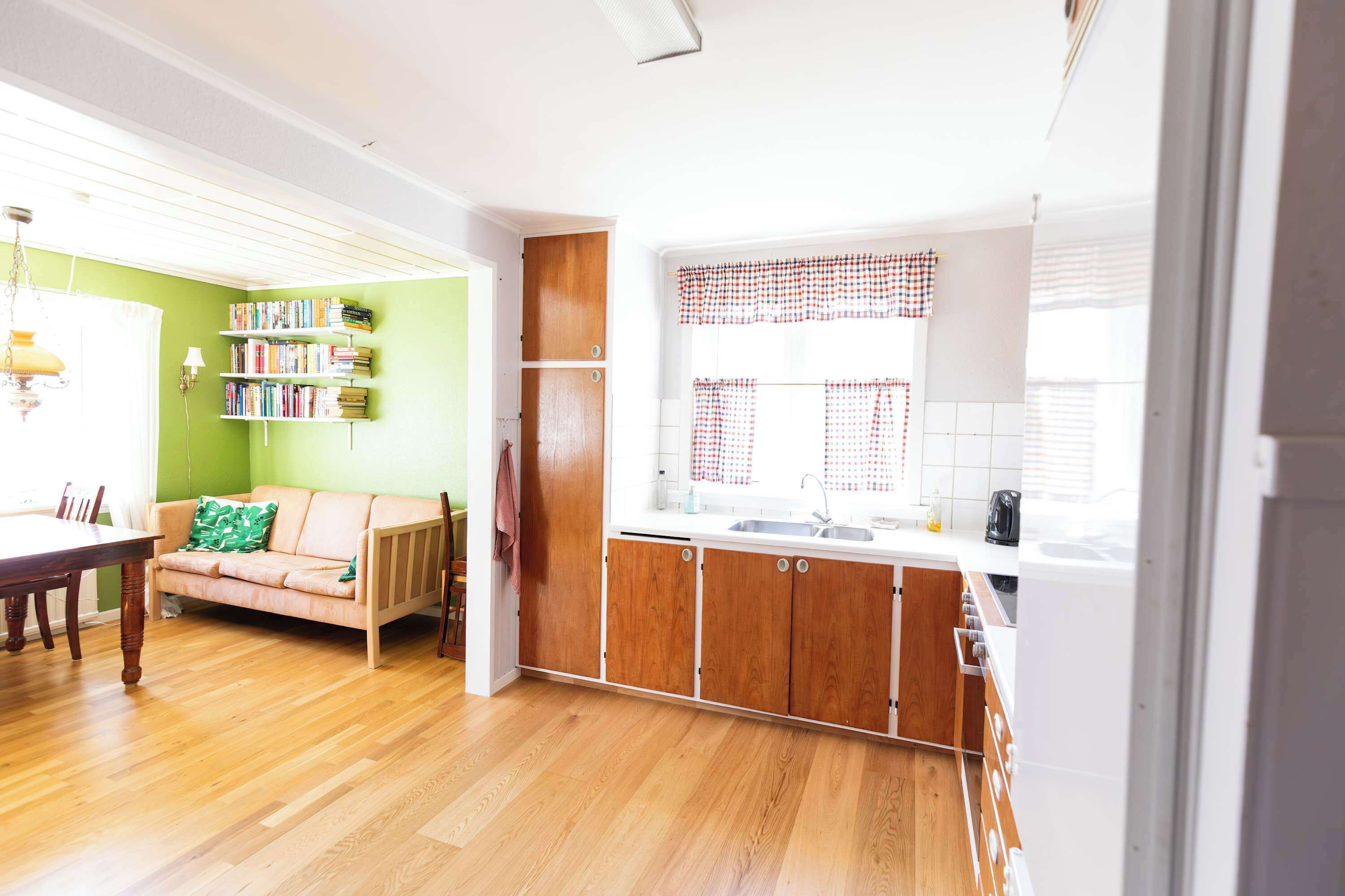 Una sala de estar luminosa con sofá, una mesa de comedor y una zona de cocina con armarios de madera y una ventana con cortinas a cuadros.
